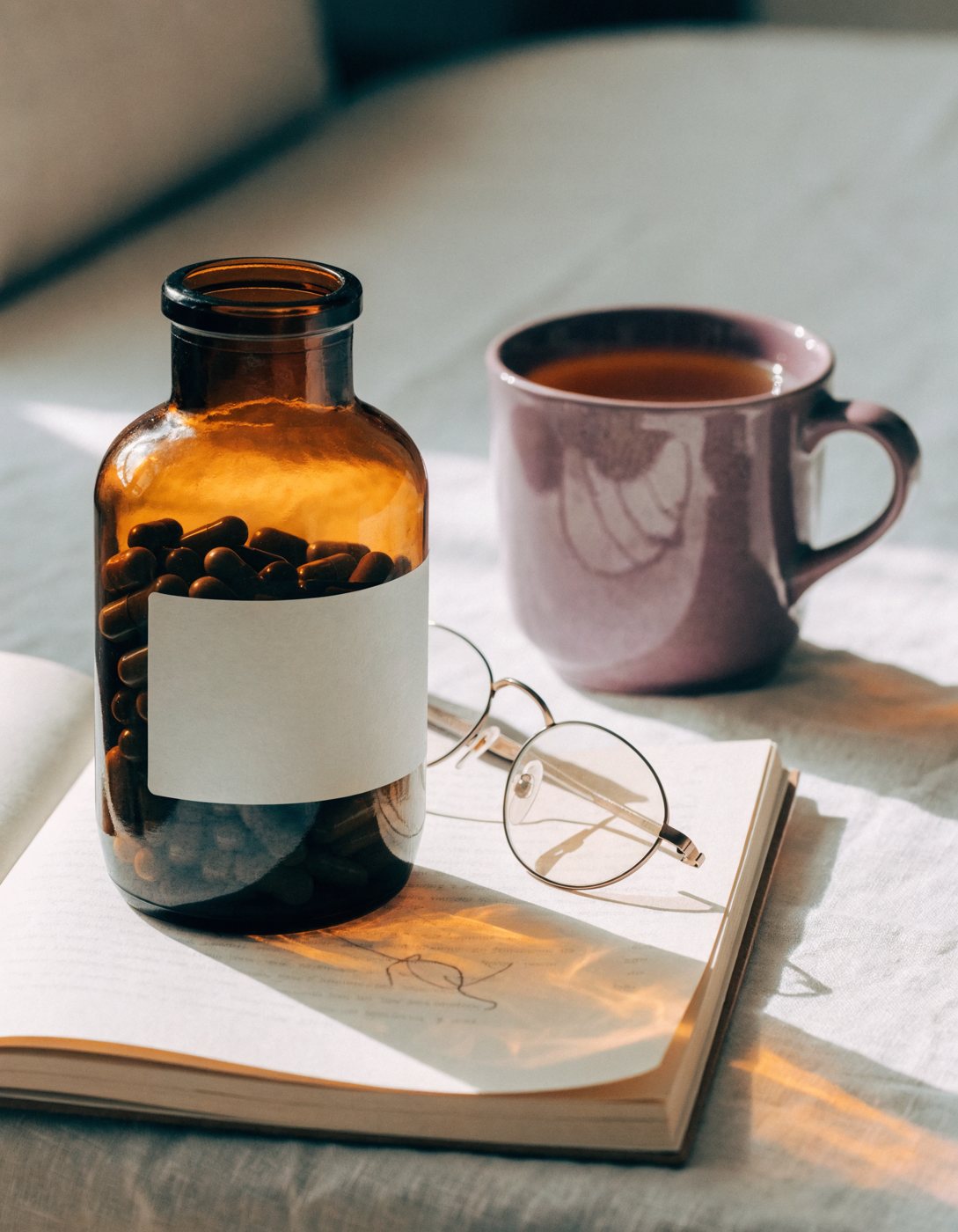 An amber apothecary jar of capsules with a blank white label, standing on an open notebook beside round reading glasses and a plum stoneware mug of tea, low sun raking across a pale linen cloth.